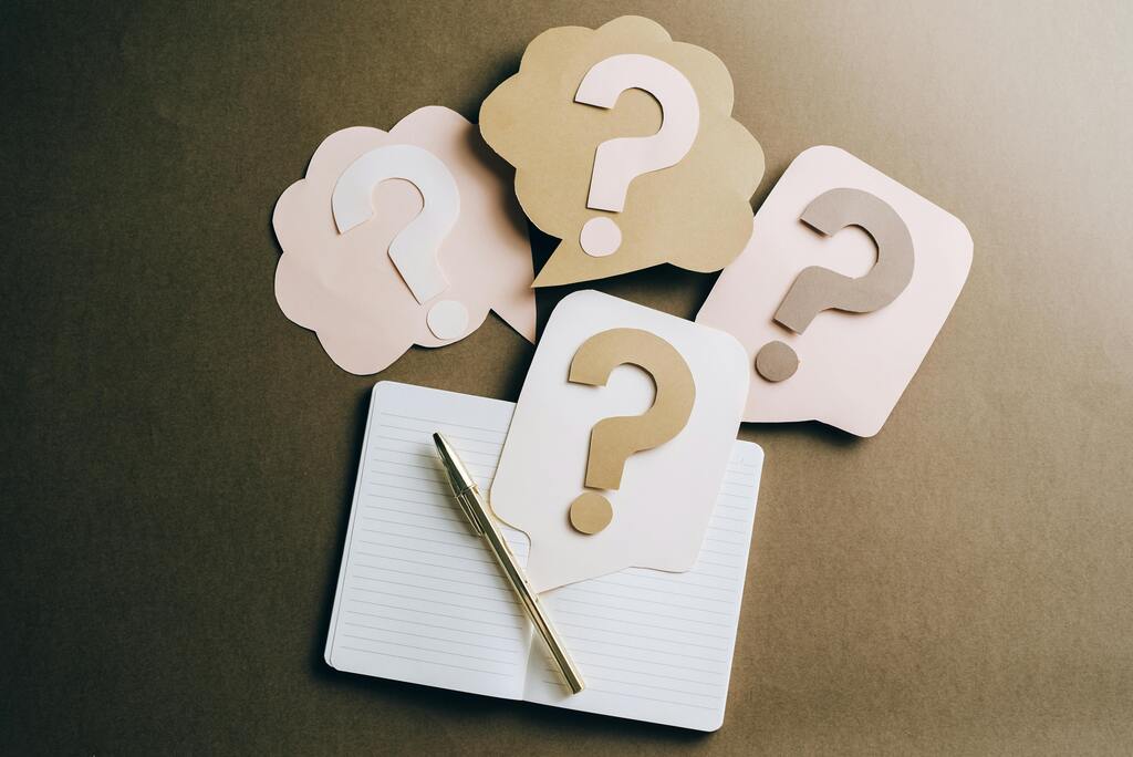 An image of blank notepaper and a pen with several sticky notes with question marks around it